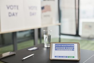 Tablet com sistema digital de registro de eleitores em uma seção eleitoral, ao lado de avisos sobre o dia da votação.