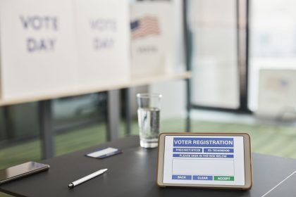 Tablet com sistema digital de registro de eleitores em uma seção eleitoral, ao lado de avisos sobre o dia da votação.