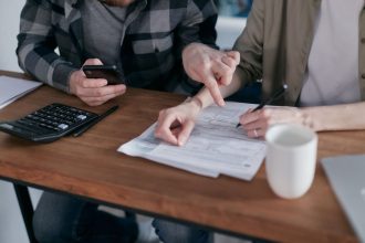 Casal analisa documentos financeiros e usa uma calculadora à mesa de casa.