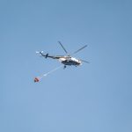 Helicóptero equipado com bolsa de água sobrevoa uma área atingida por incêndio florestal.