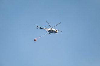 Helicóptero equipado com bolsa de água sobrevoa uma área atingida por incêndio florestal.