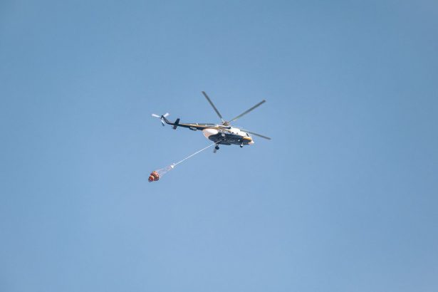 Helicóptero equipado com bolsa de água sobrevoa uma área atingida por incêndio florestal.